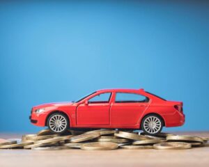 A red toy car sits atop a stack of gold coins against a blue background, symbolizing wealth and investment in automobiles.