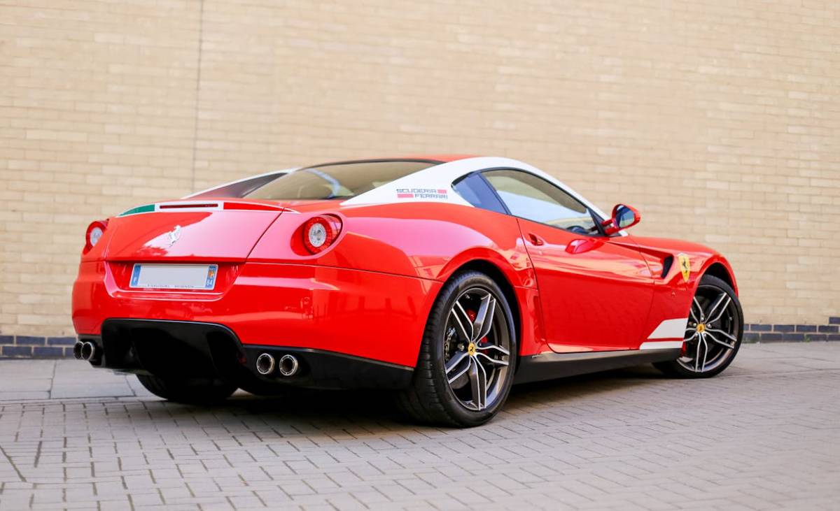 A sleek red Ferrari with a white roof, featuring sporty black wheels and exhaust, parked against a light brick wall.