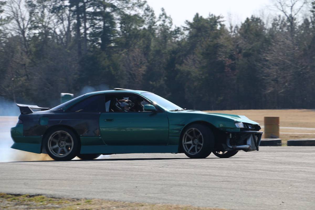 A teal and black sports car drifts on a track, with smoke trailing behind and a dog visible in the driver's seat.