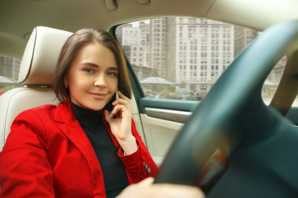 A person sitting in a car, wearing a red jacket, holding a phone, with city buildings visible through the windshield.