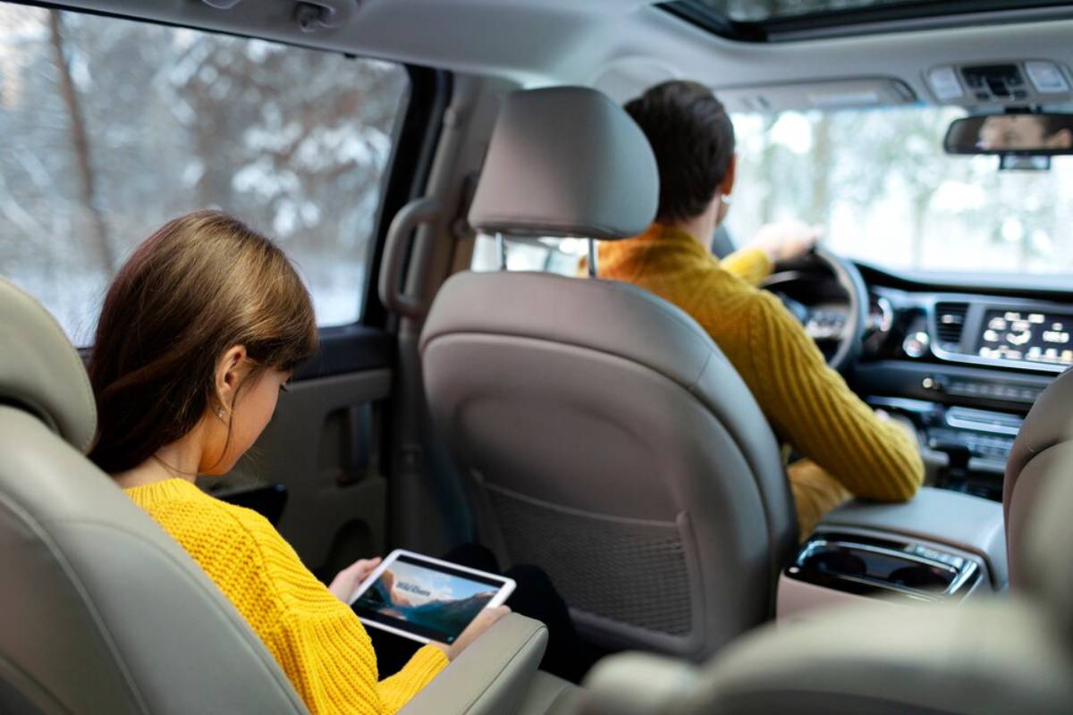 A child wearing a yellow sweater sits in a car, focused on a tablet, while an adult drives in the front seat.