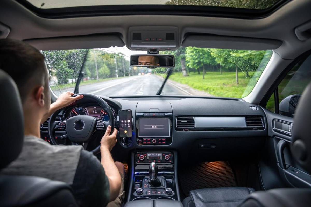 A person driving a car with a steering wheel in one hand and a phone in the other, rainy weather outside.