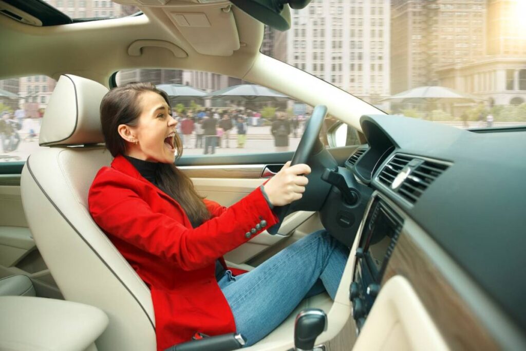 A woman in a red blazer drives a modern car, with a cityscape visible through the windshield.