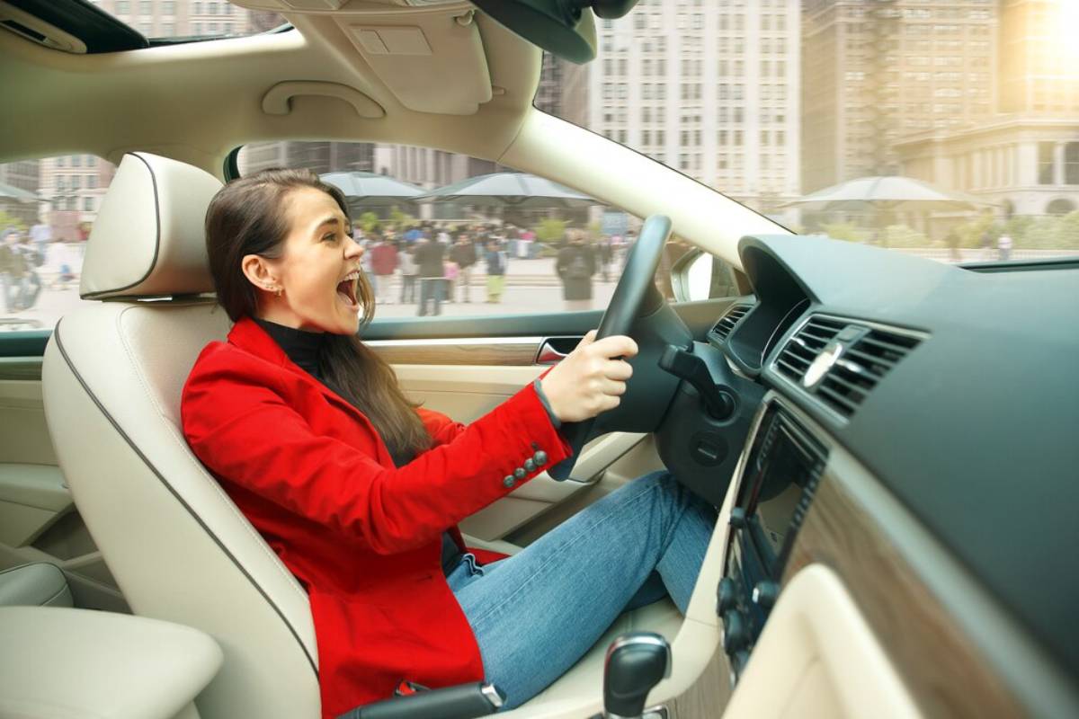 A woman in a red blazer drives a modern car, with a cityscape visible through the windshield.