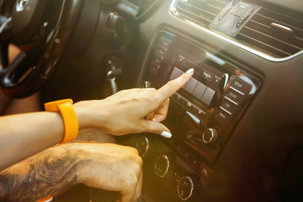 A hand with a bright orange smartwatch interacts with a car's touchscreen, selecting the login option.