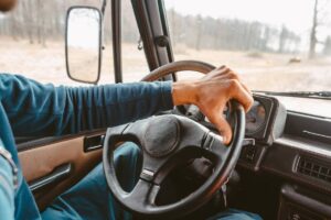 A person gripping a steering wheel inside a vehicle, with trees and a blurred landscape visible through the windshield.
