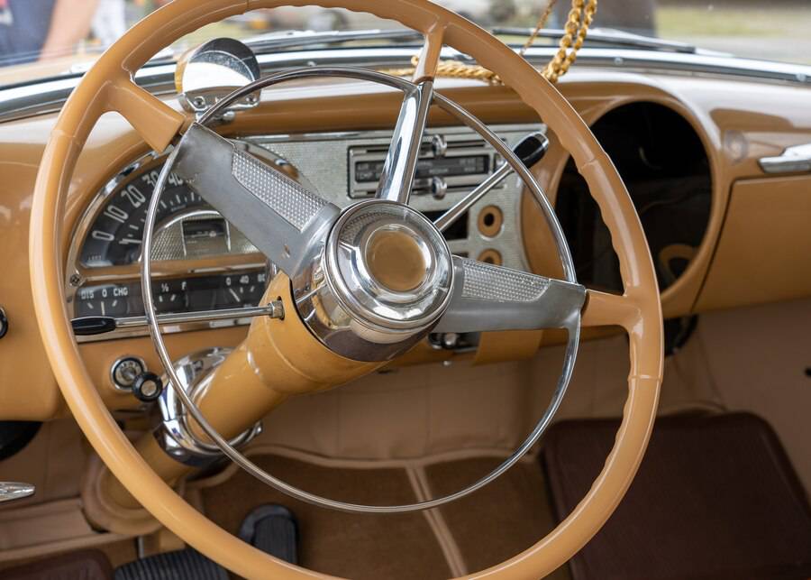Close-up of a vintage car's dashboard featuring a classic chrome steering wheel and retro control panel in warm beige tones.