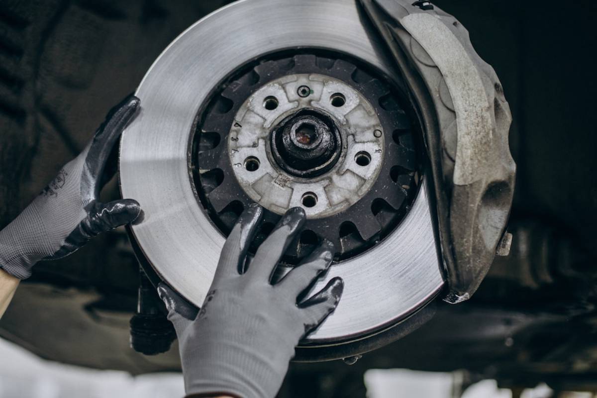 A mechanic's hands in gray gloves hold a shiny brake disc while working on a car, focused on vehicle maintenance.