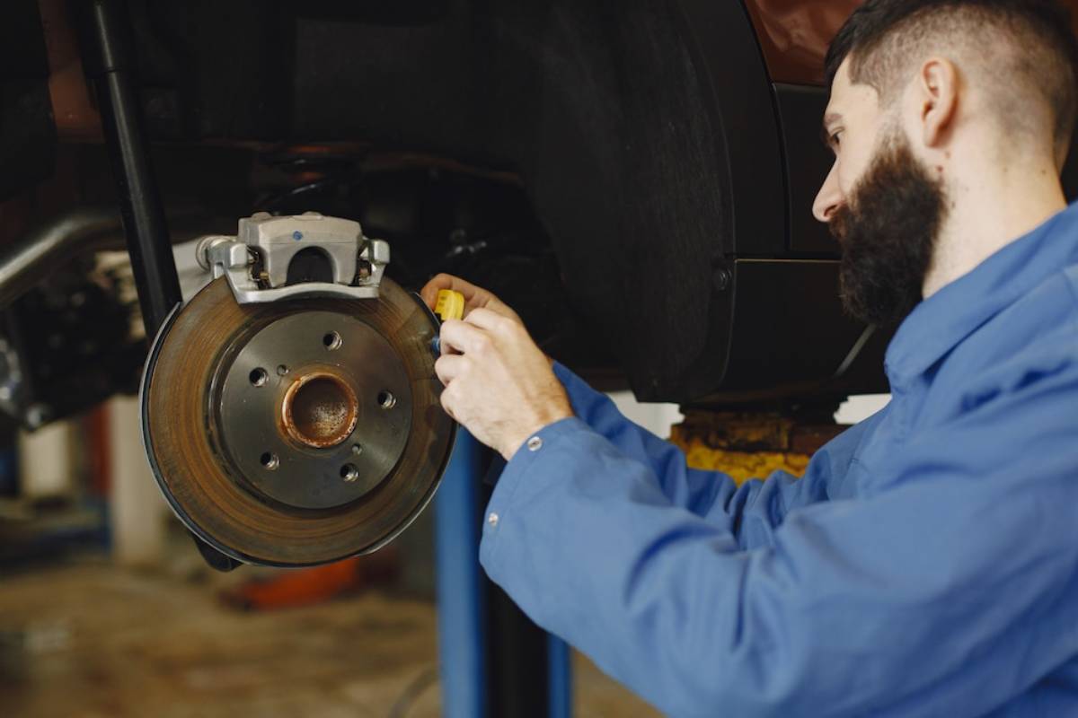 A mechanic in a blue shirt works on a car's brake system, holding a yellow tool near the brake disc and caliper.