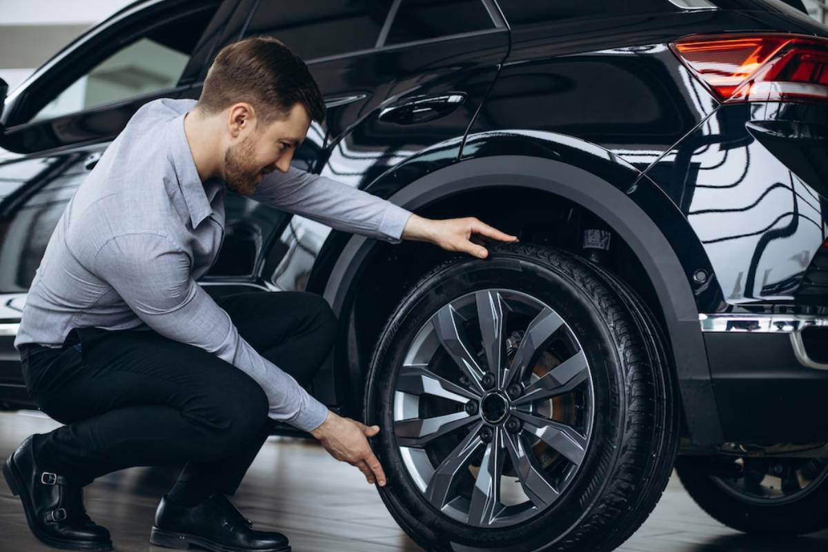 A man in a light shirt inspects the tire of a sleek black SUV, focusing on its rim and tread in a well-lit automotive showroom.