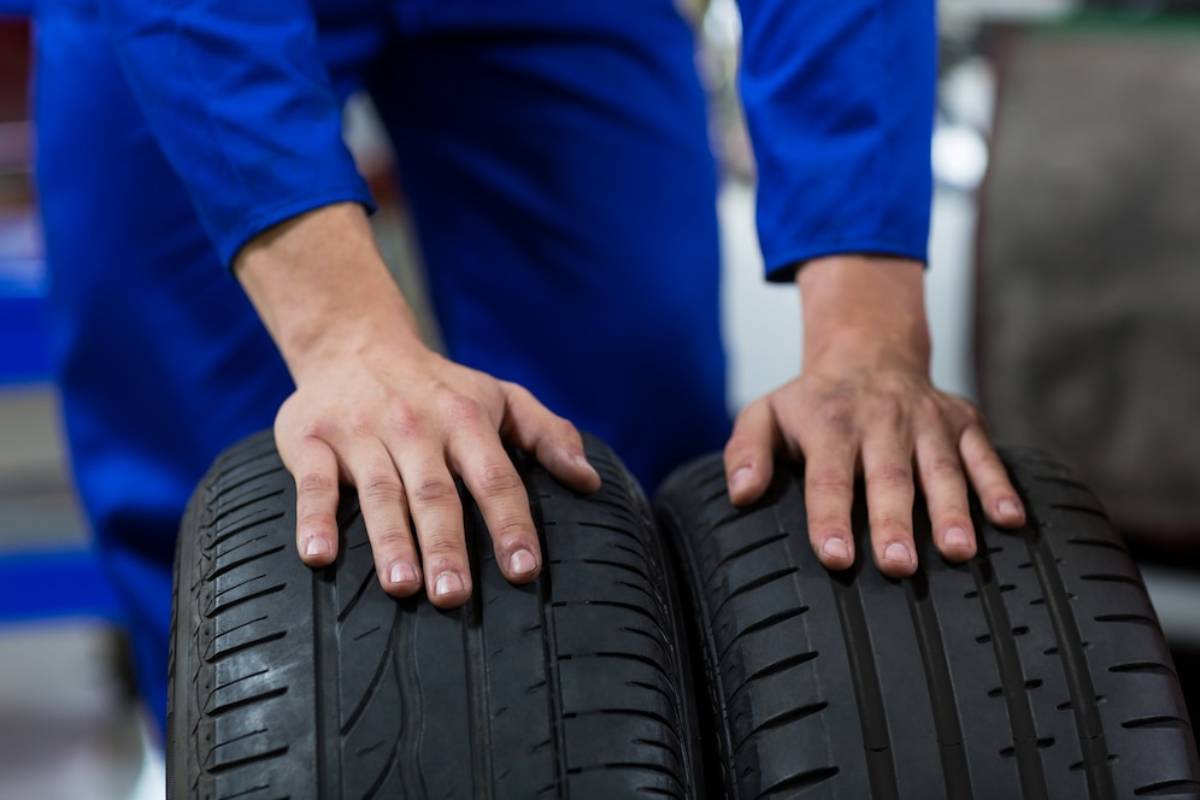 A mechanic in a blue jumpsuit examines two car tires, placing hands on the treads for inspection in a workshop setting.
