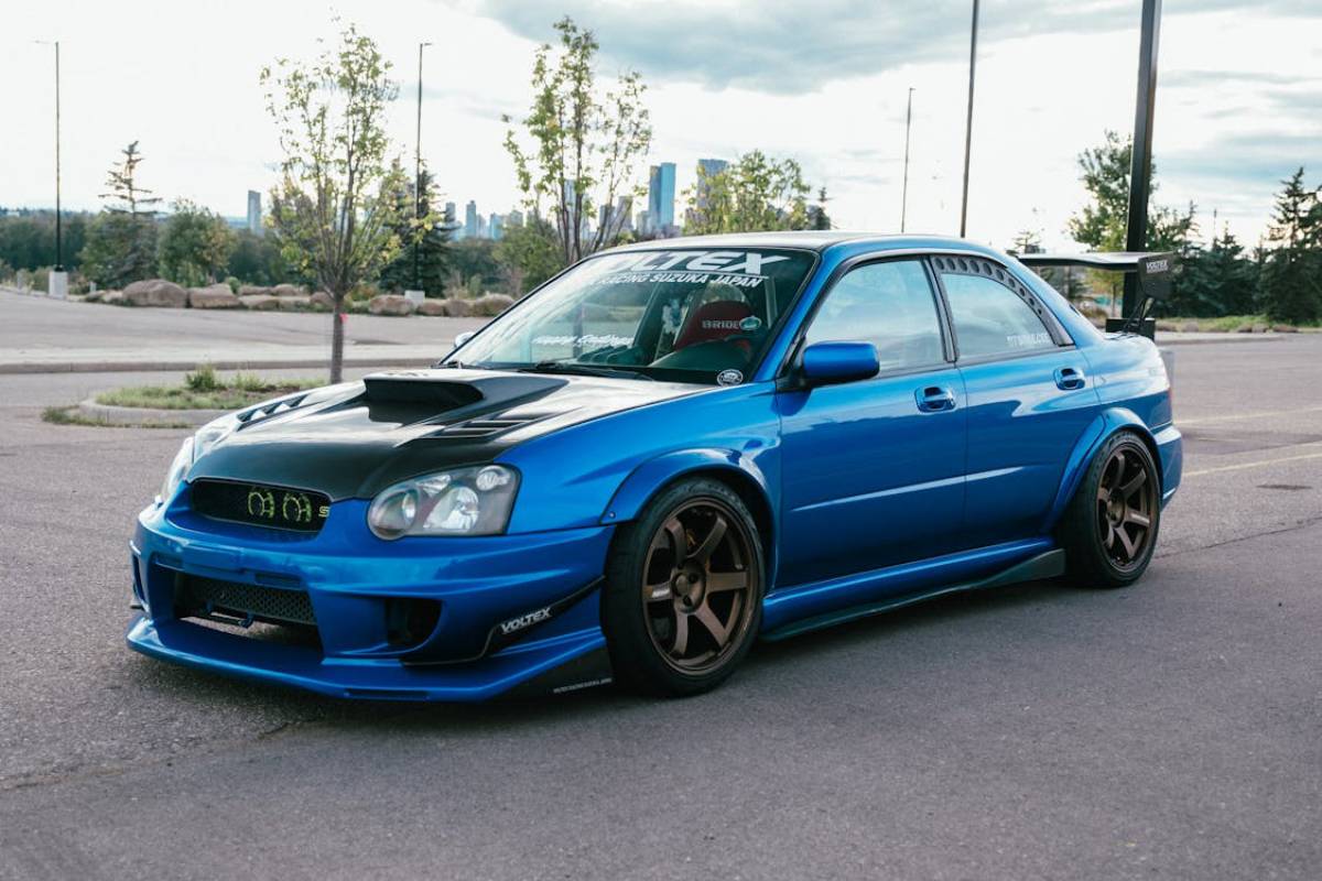 A modified blue Subaru Impreza with a black hood and bronze rims parked in a scenic area.
