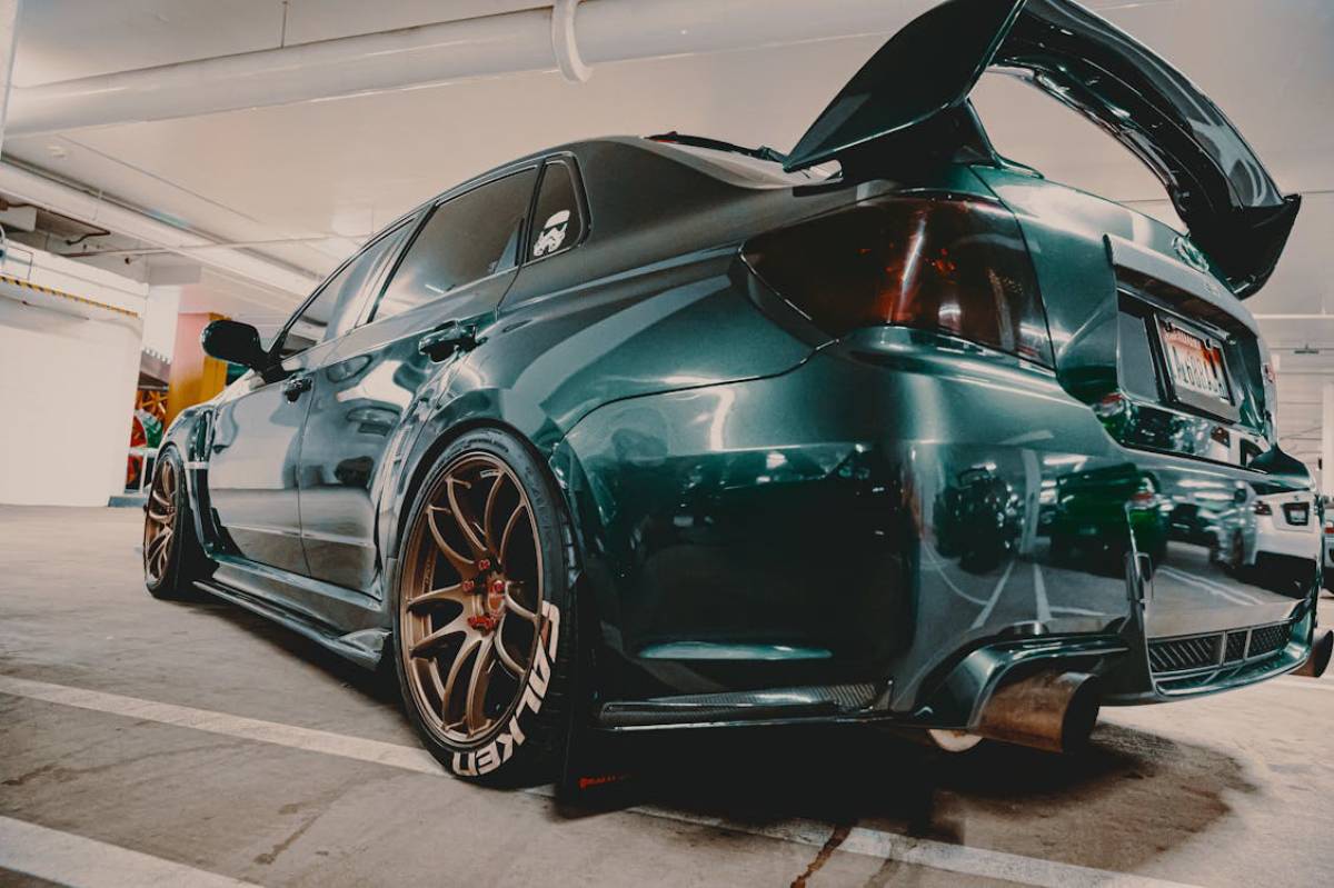 A modified green sedan with a large rear spoiler and bronze wheels parked in an underground garage.