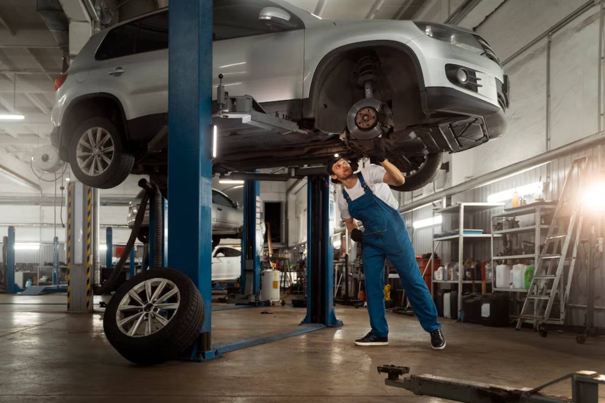 A mechanic in blue overalls works underneath a silver SUV elevated on a lift in a well-equipped garage.