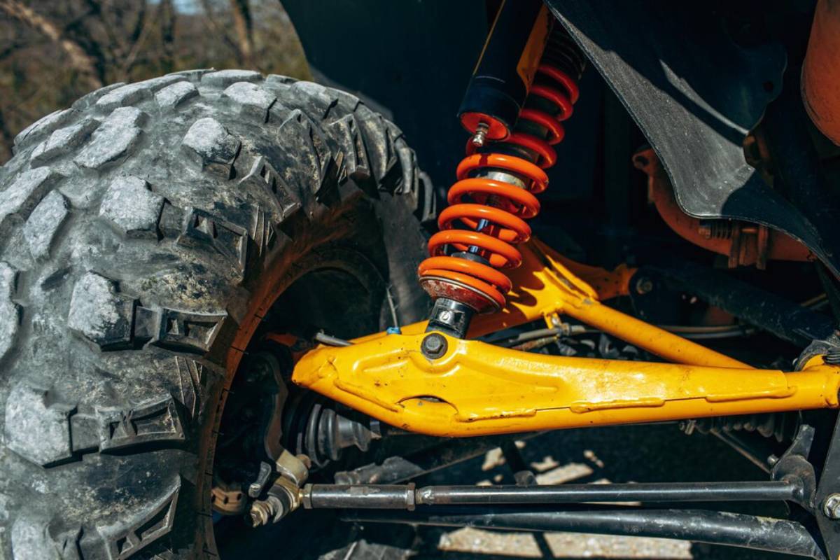 Close-up of a rugged off-road vehicle's suspension, featuring an orange coil spring and a yellow control arm next to a large, textured tire.