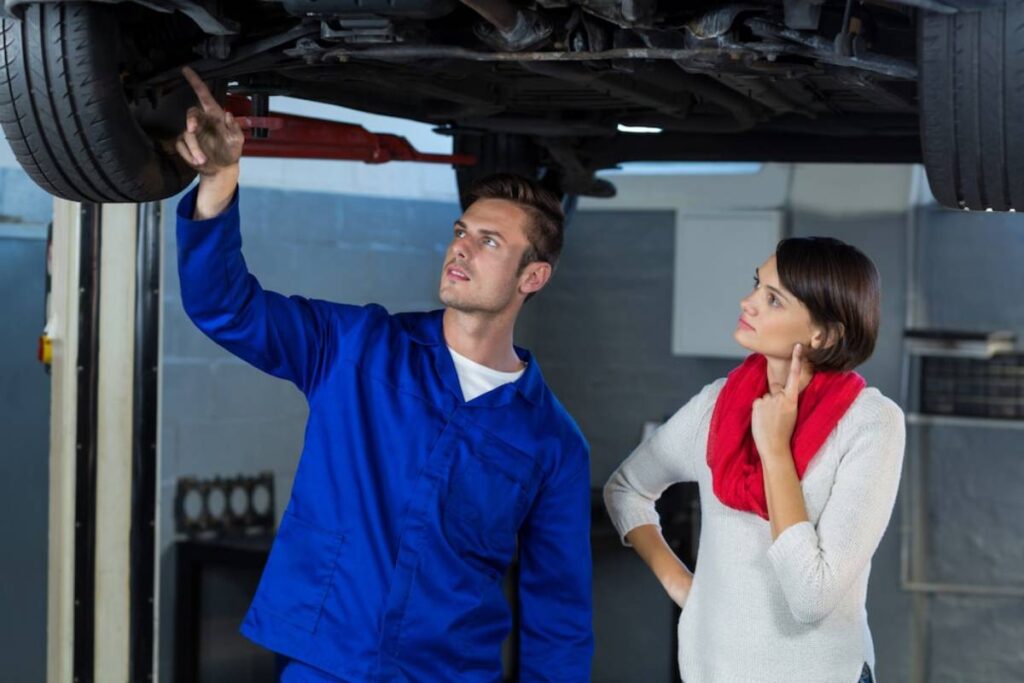 A mechanic in a blue uniform points to an elevated vehicle's undercarriage while a woman in a red scarf listens attentively.