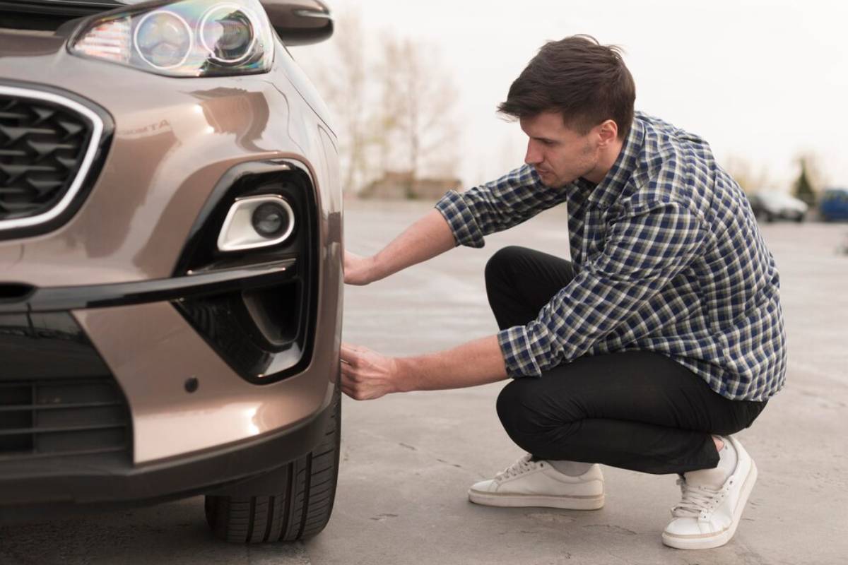 A person crouches next to a brown car, inspecting its tire, wearing a plaid shirt and white sneakers in an outdoor setting.