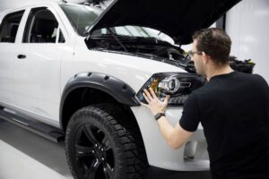 A person inspects the front of a white SUV with an open hood, focusing on the headlights and fender details.