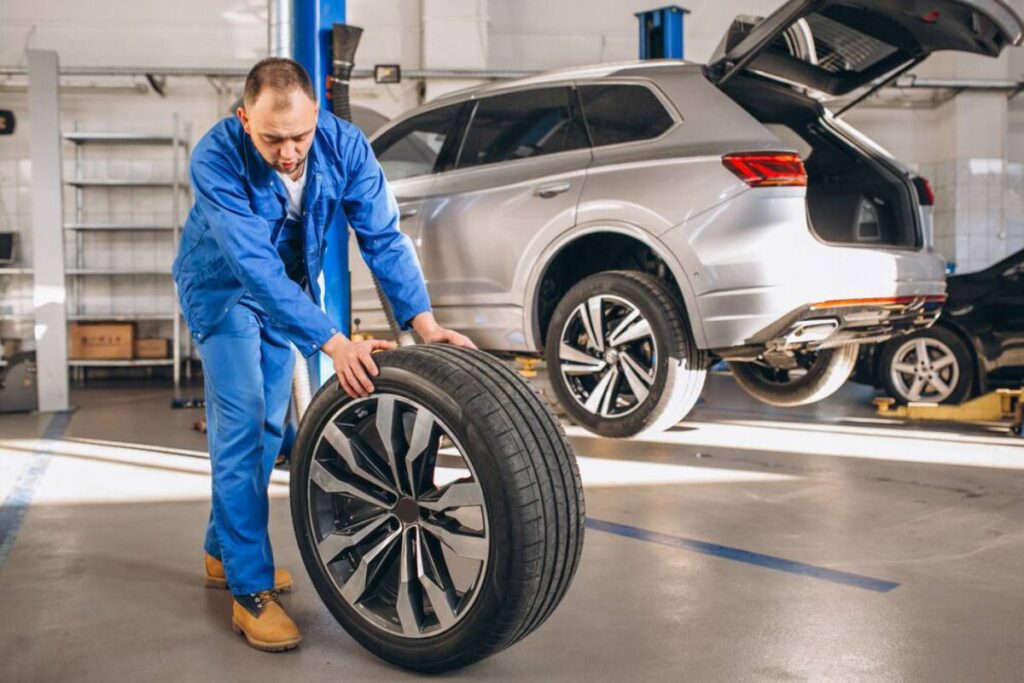 A mechanic in blue overalls rolls a tire in an auto repair shop, with a silver SUV on a lift in the background.