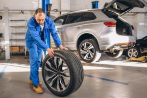 A mechanic in blue overalls rolls a tire in an auto repair shop, with a silver SUV on a lift in the background.