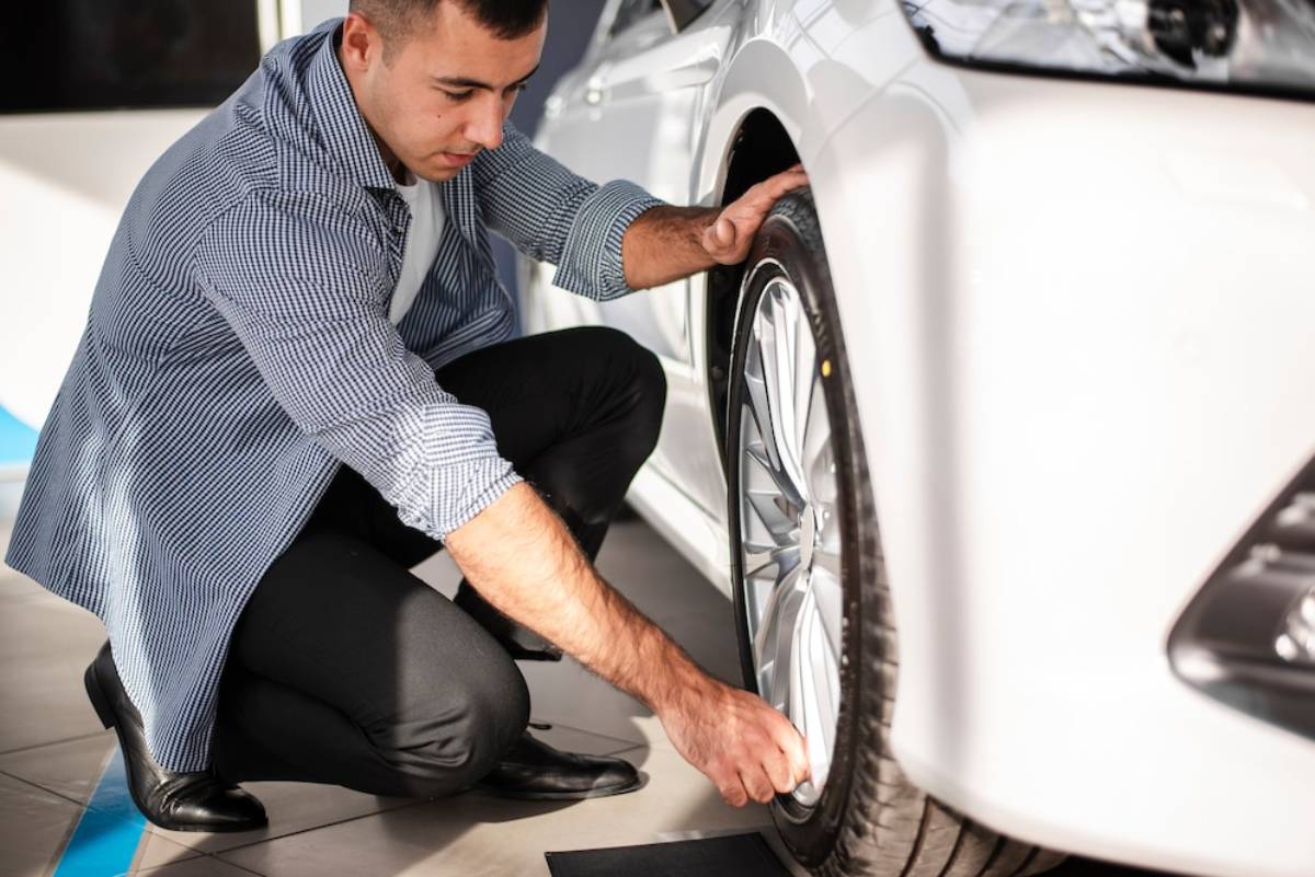 A person kneels beside a white car, examining its tire with a focused expression.