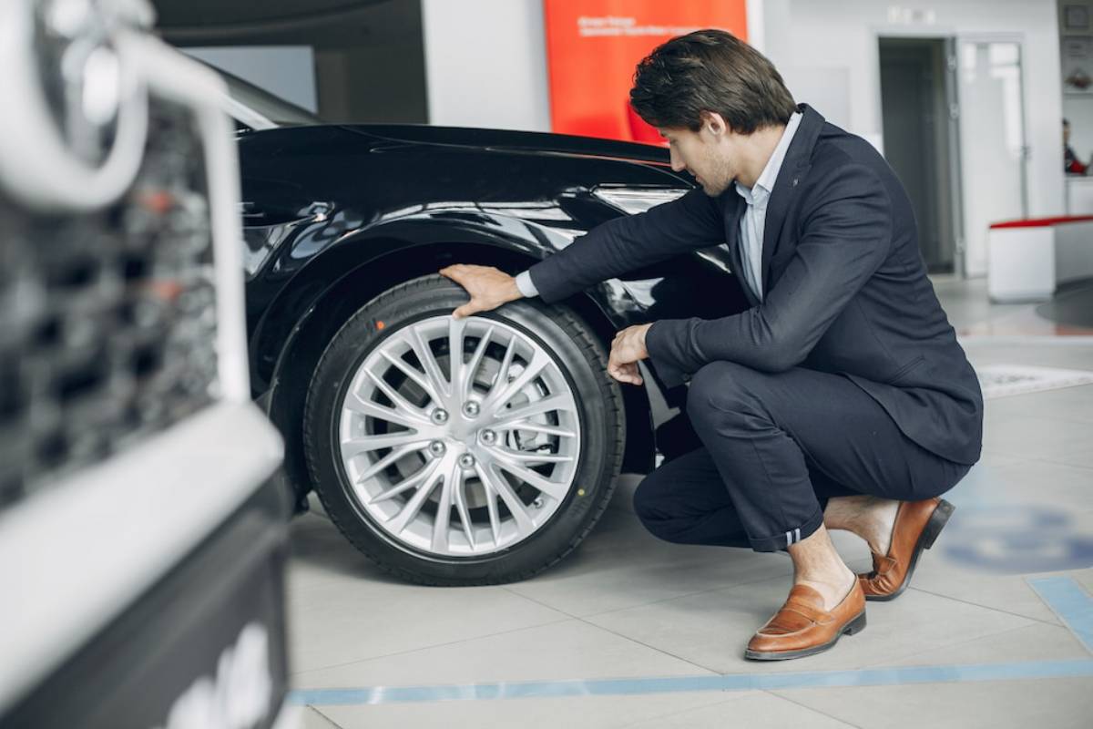 A man in a formal suit kneels beside a sleek black car, inspecting its wheel attentively in a modern showroom environment.
