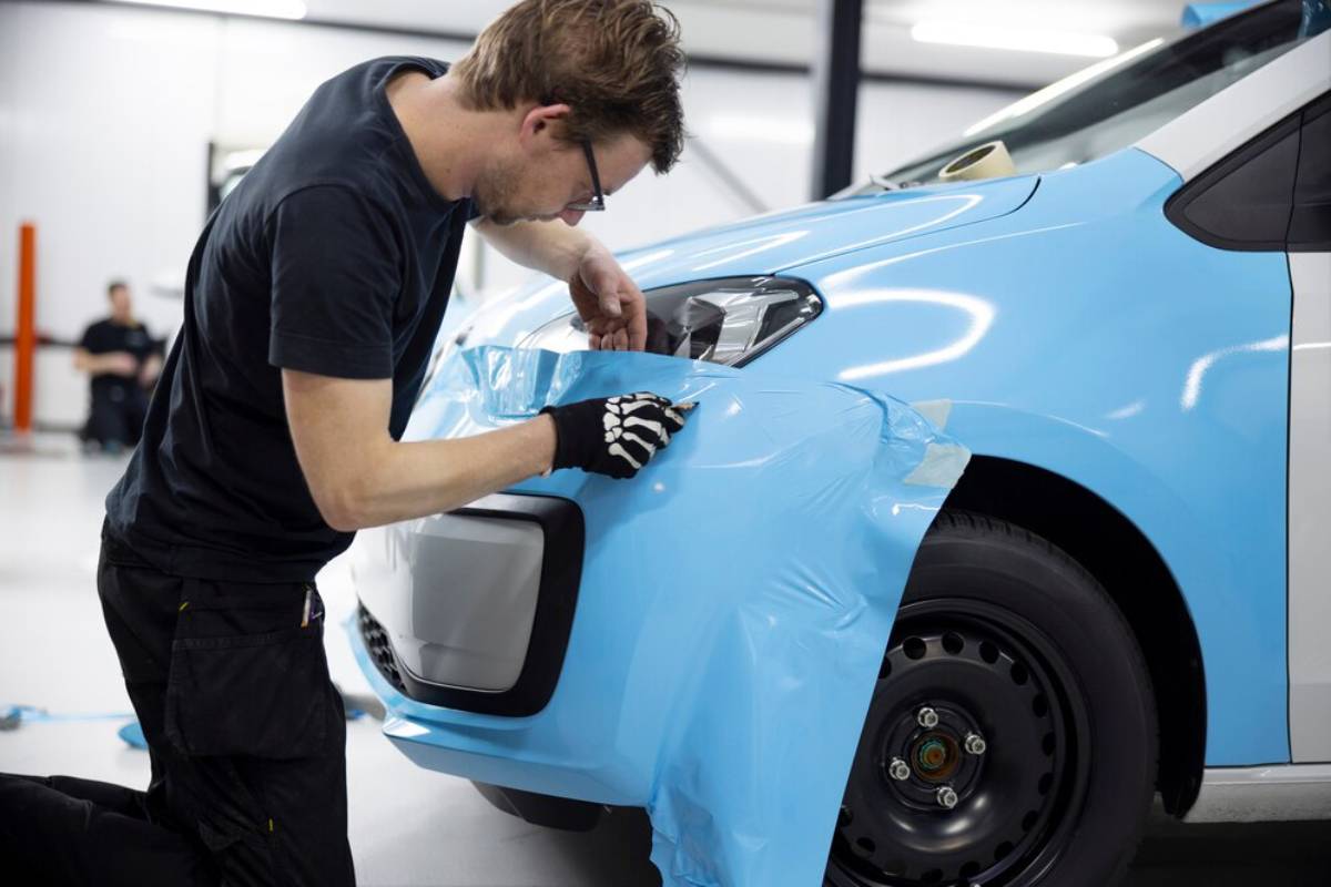 A person applies a blue vinyl wrap to the front of a vehicle in a workshop, focusing on smoothing out the material.