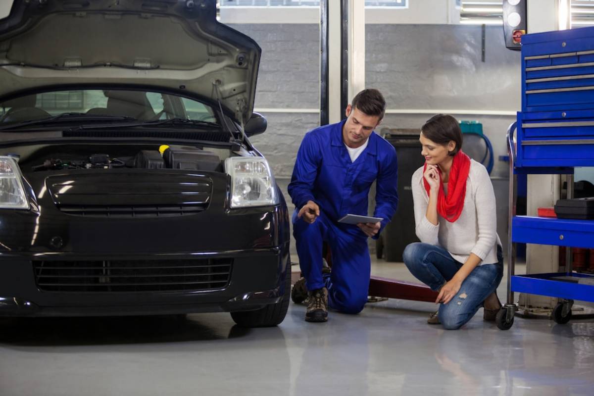 A mechanic discusses car maintenance with a female client, both focused on the vehicle's open hood in a well-equipped garage.