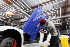 A technician applies a blue vinyl wrap to the door of a white sports car in a modern workshop with neon lights.