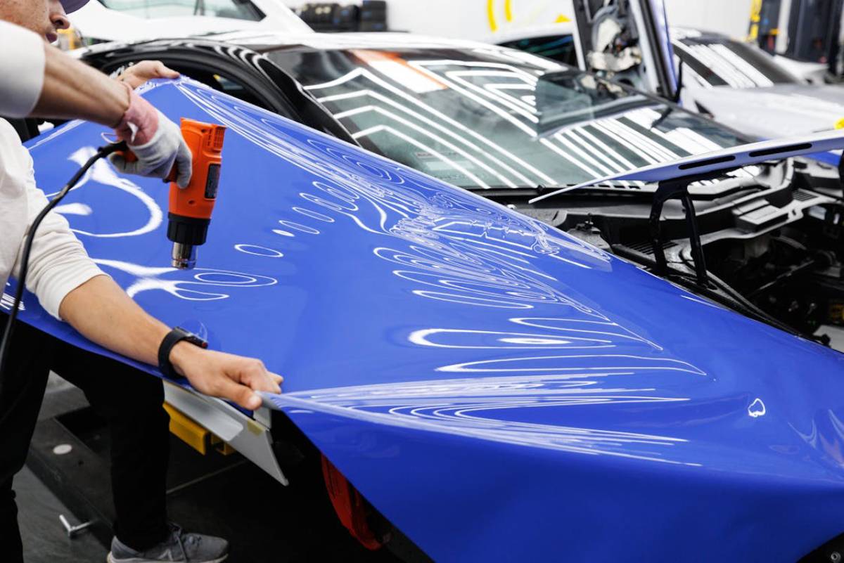 A person using a heat gun to apply a reflective blue vinyl wrap to a car's exterior in a workshop setting.