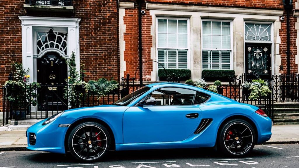 A vibrant blue Porsche Cayman parked in front of a historic red brick building with large windows and a decorative black door.