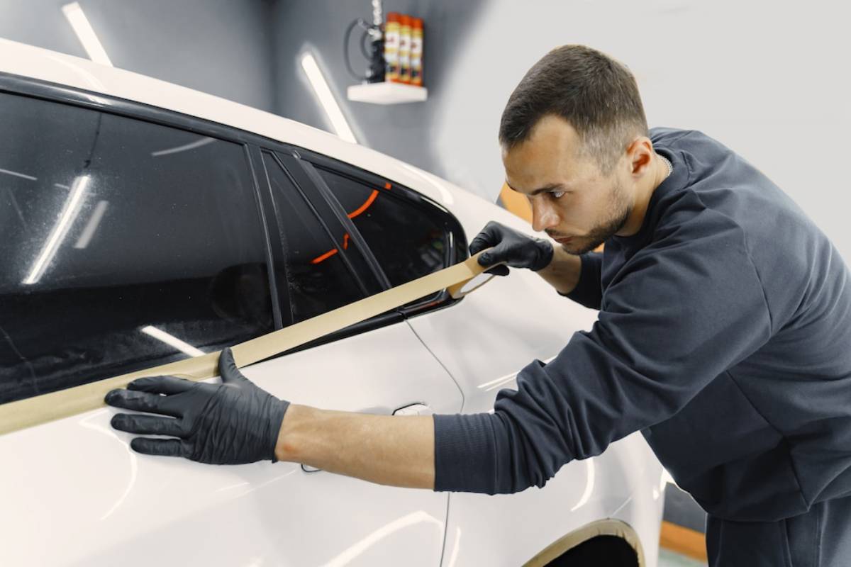 A technician wearing gloves carefully applies tape along the edge of a white car's window.
