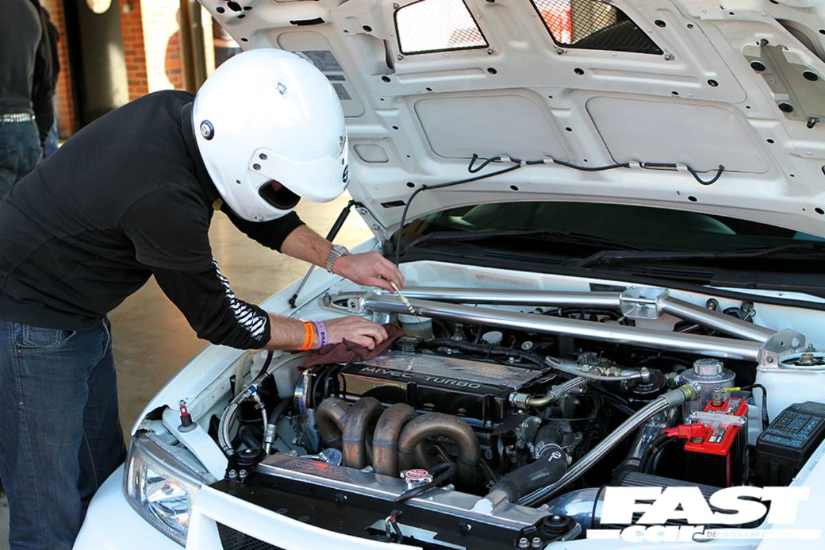 A mechanic in a racing helmet works on a turbocharged engine, leaning over the open hood of a white car in a garage setting.