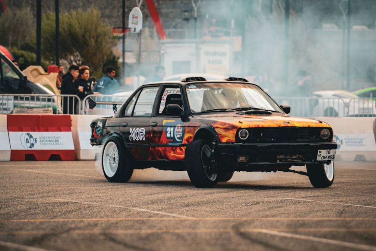 A modified car with a fiery design performs a drift on a racetrack, surrounded by spectators and smoke from the tires.