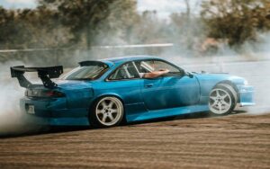 A blue sports car performs a drift on a dusty track, surrounded by billowing smoke and trees in the background.