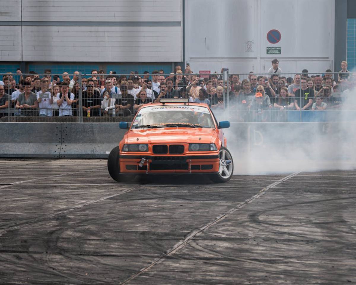 An orange drifting car generates smoke as it skids on a track, while a crowd of spectators watches excitedly in the background.