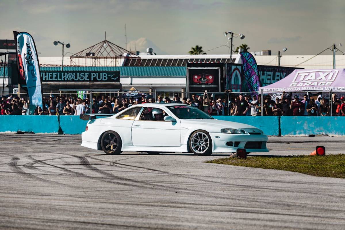 A white sports car drifts on a racetrack, surrounded by an enthusiastic crowd at a motorsport event.