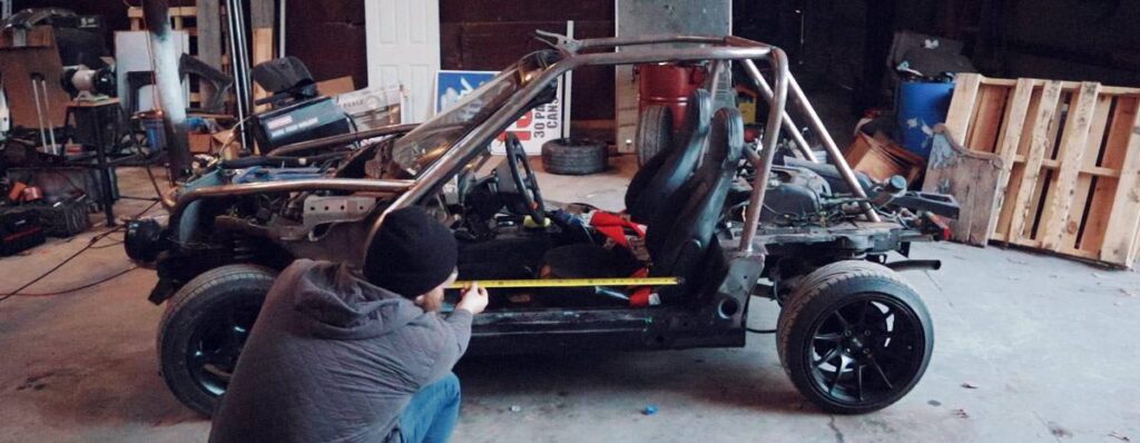 A person measures a partially assembled race car chassis in a cluttered garage.