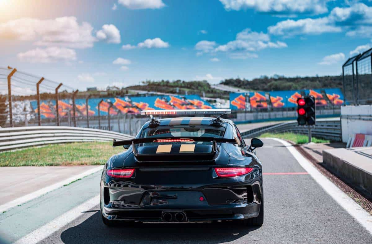 A black racing car with a large rear wing and lights, waiting at a red traffic signal on a racetrack under a blue sky.