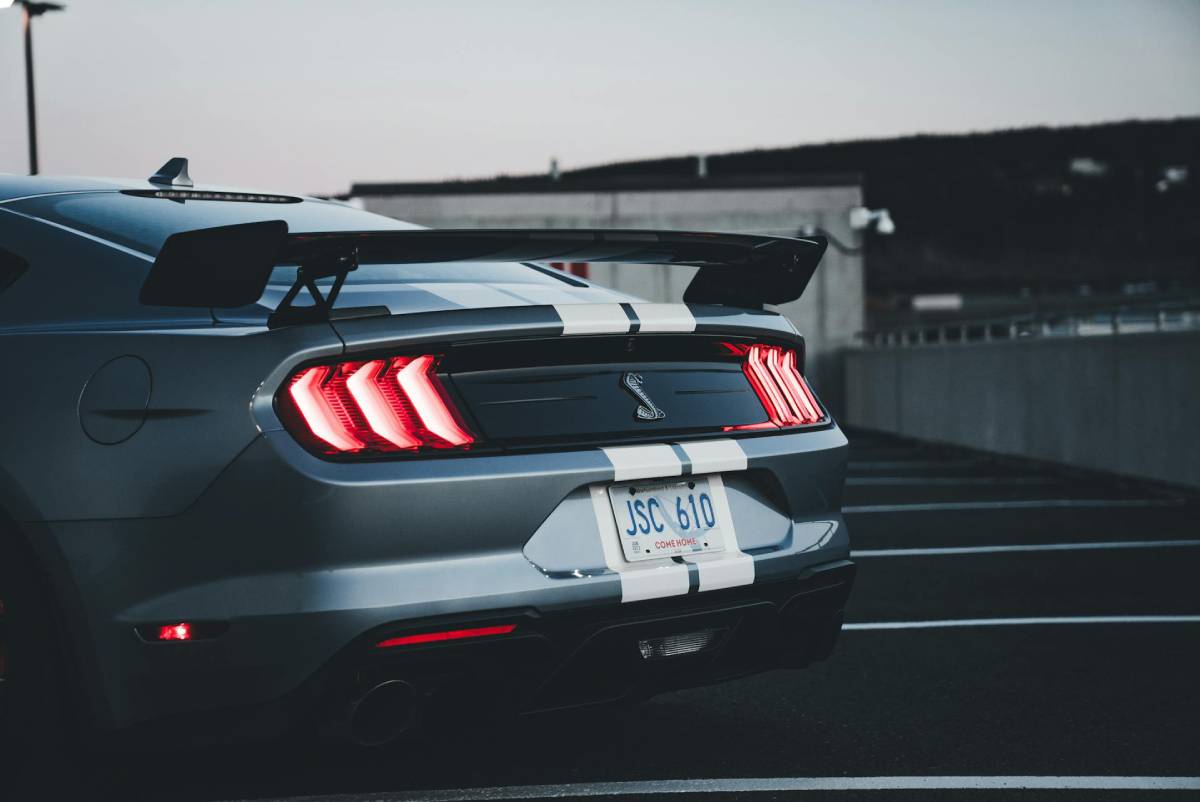 A sleek gray Shelby Mustang with a rear spoiler, featuring iconic striped design and glowing taillights, parked in a dimly lit area.