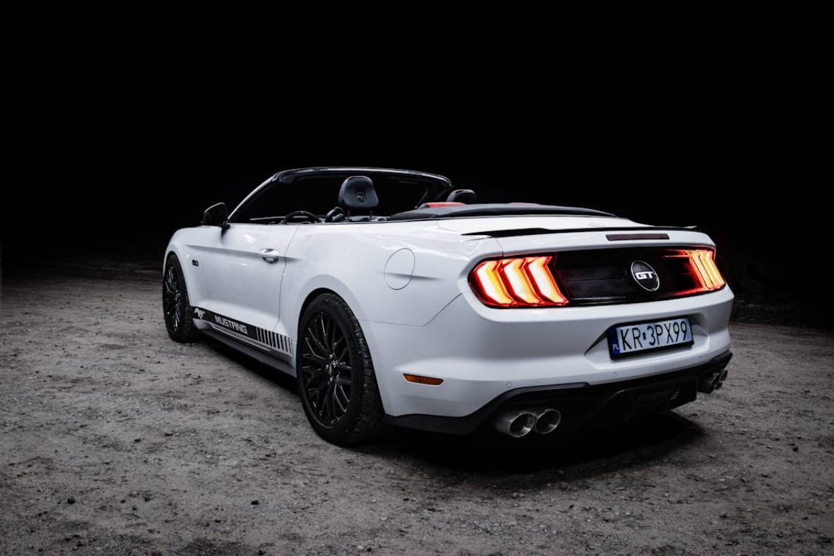 A sleek white Ford Mustang GT convertible parked on gravel at night, showcasing its bold rear lights and sporty design.