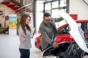 A woman consults with a man at a car dealership while he inspects the engine of a vehicle.