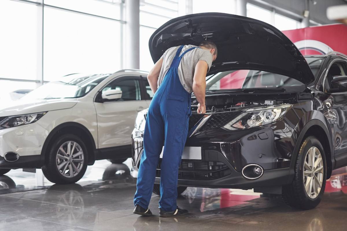 A mechanic in blue overalls inspects the engine of a black car in a bright dealership, with a white vehicle visible nearby.