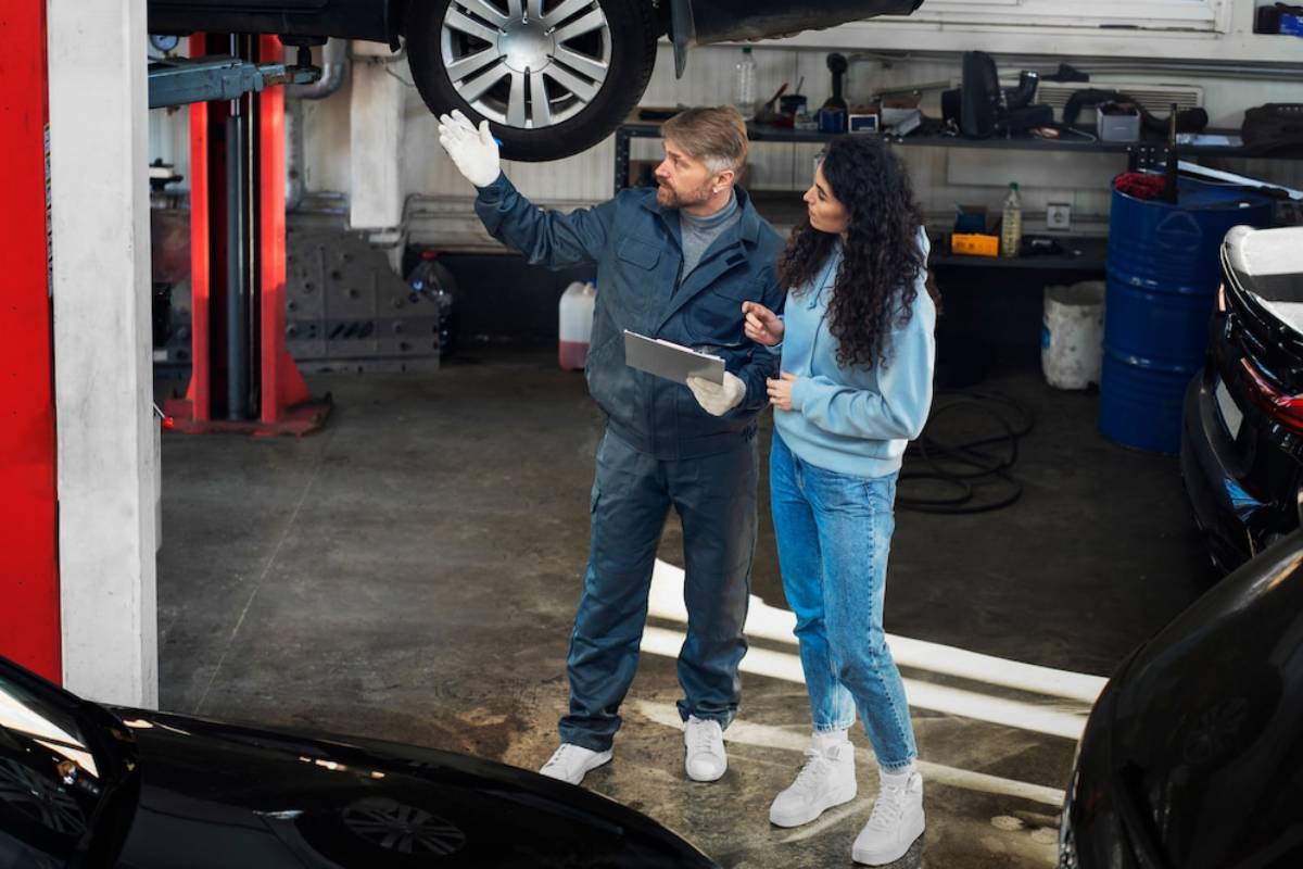 A mechanic gestures while explaining something to a customer in a garage, with cars and tools visible in the background.