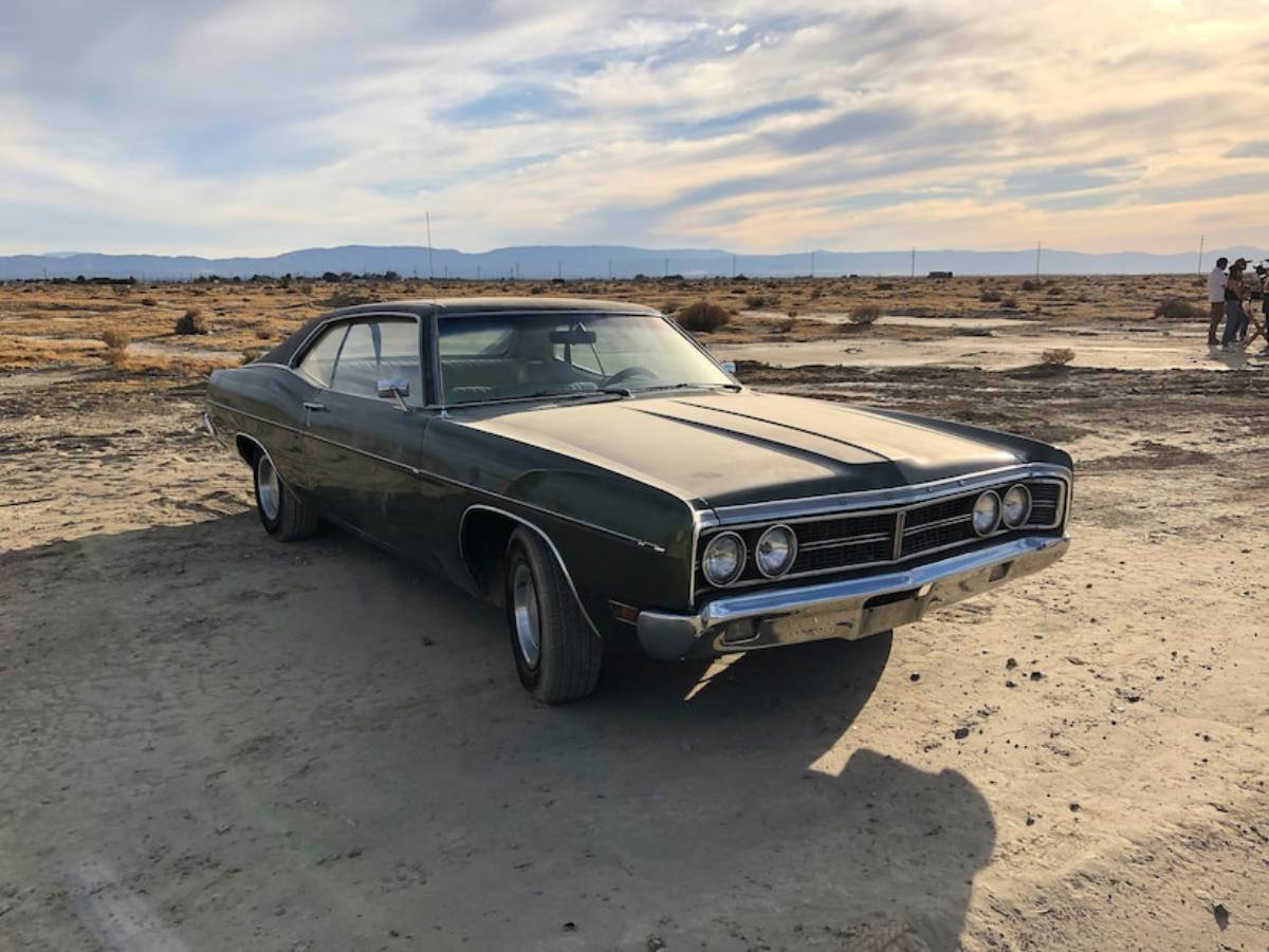 A vintage green muscle car parked in a desolate, arid landscape under a cloudy sky.