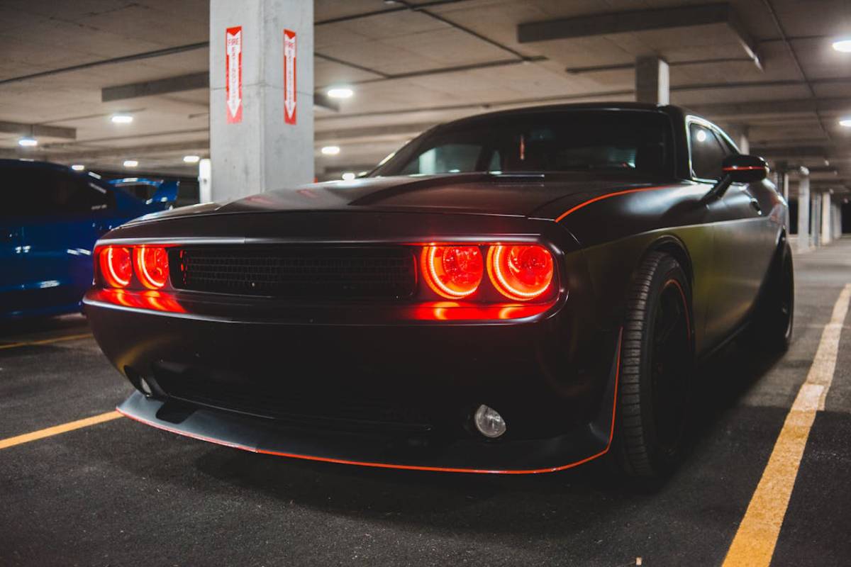 A sleek black muscle car with red glowing headlights, parked in a dimly lit underground garage alongside a blue car.