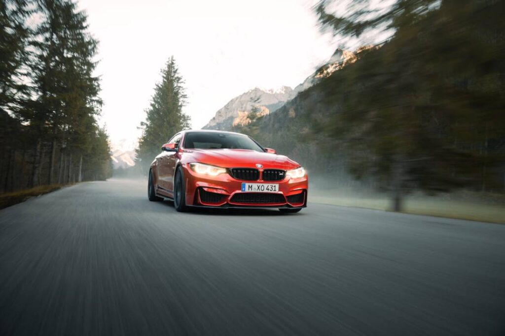 A bright red BMW M4 speeds along a winding mountain road, flanked by tall pine trees and misty mountains in the background.