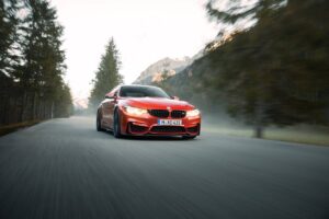 A bright red BMW M4 speeds along a winding mountain road, flanked by tall pine trees and misty mountains in the background.