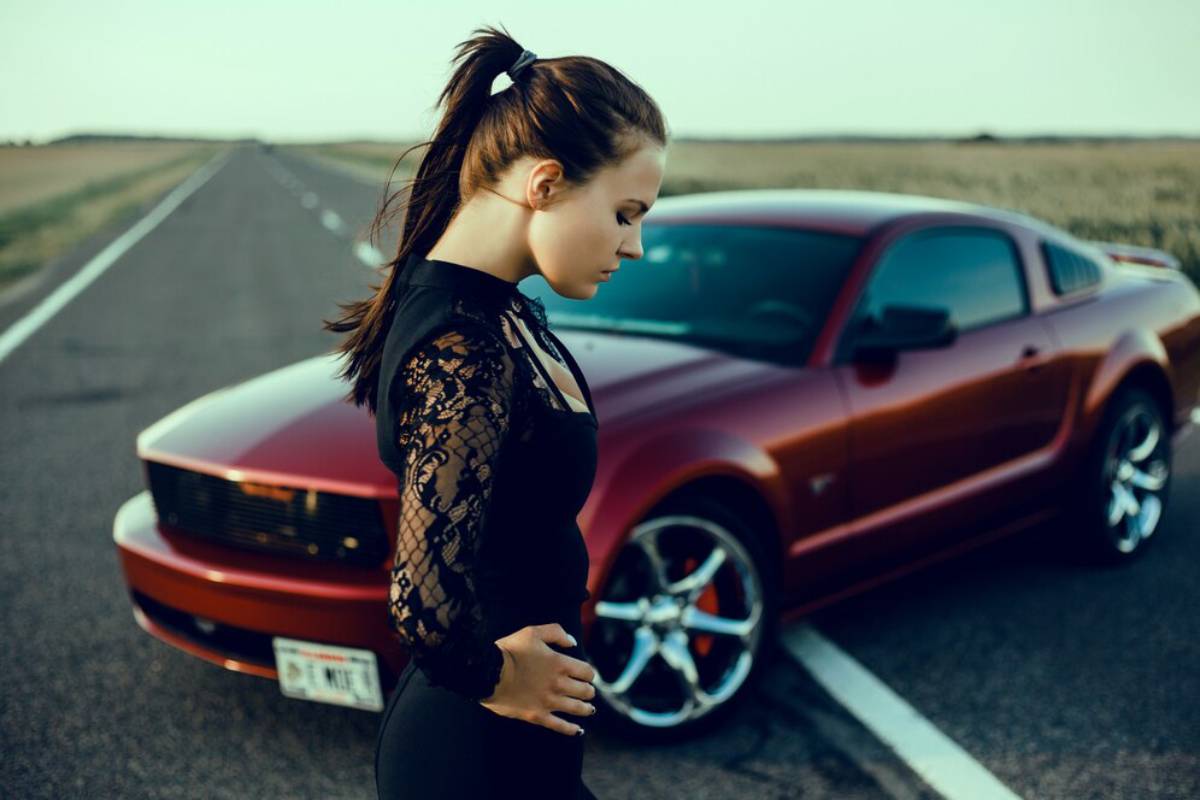 A young woman in a black lace top stands confidently beside a shiny red sports car on an open road.