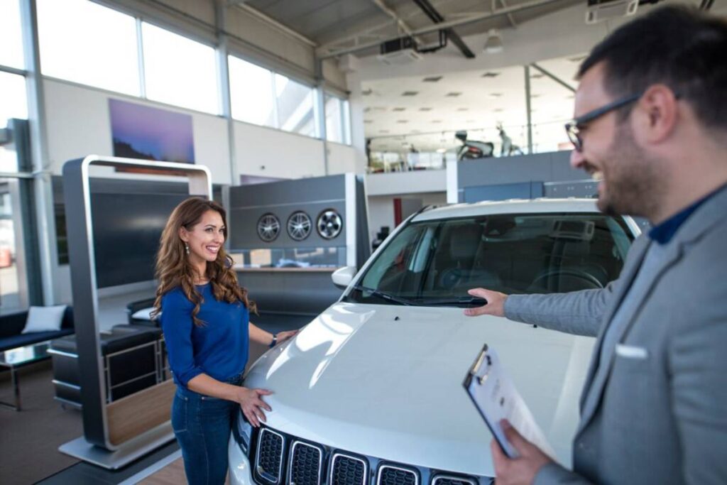 A woman in a blue shirt examines a white car in a bright, modern showroom, while a man gestures towards it with a clipboard.